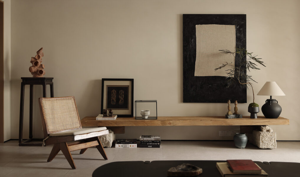 Minimalist living space with wooden bench, sculptural decor, woven lounge chair, neutral wall art, and warm natural lighting