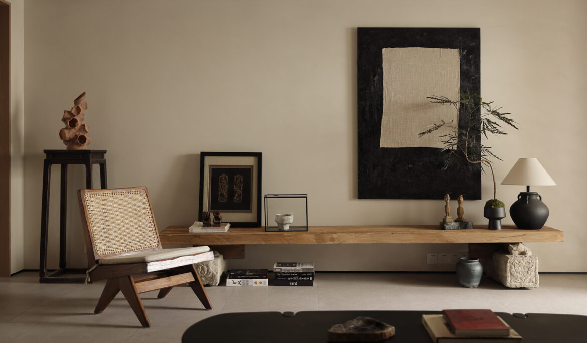 Minimalist living space with wooden bench, sculptural decor, woven lounge chair, neutral wall art, and warm natural lighting