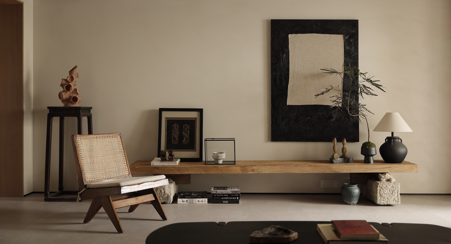 Minimalist living space with wooden bench, sculptural decor, woven lounge chair, neutral wall art, and warm natural lighting