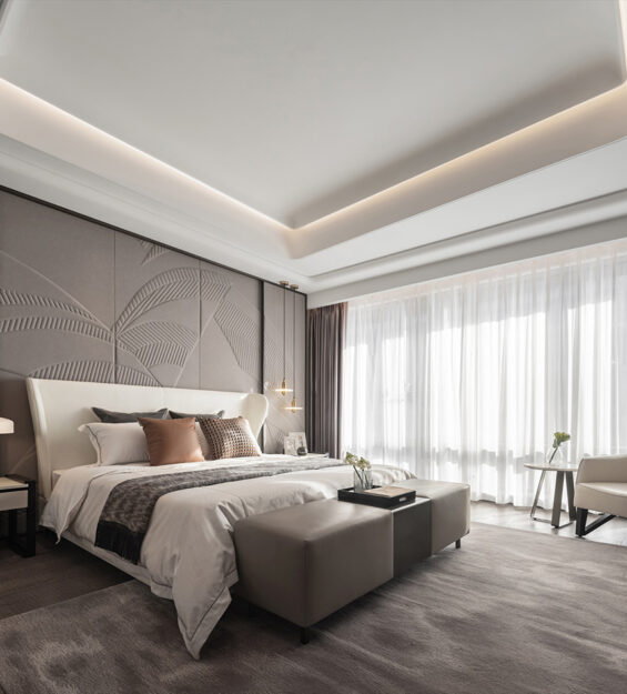 Contemporary bedroom with an upholstered feature wall, neutral‑toned bed and bench, soft cove ceiling lighting, bedside lamps, and a lounge chair beside floor‑to‑ceiling sheer curtains