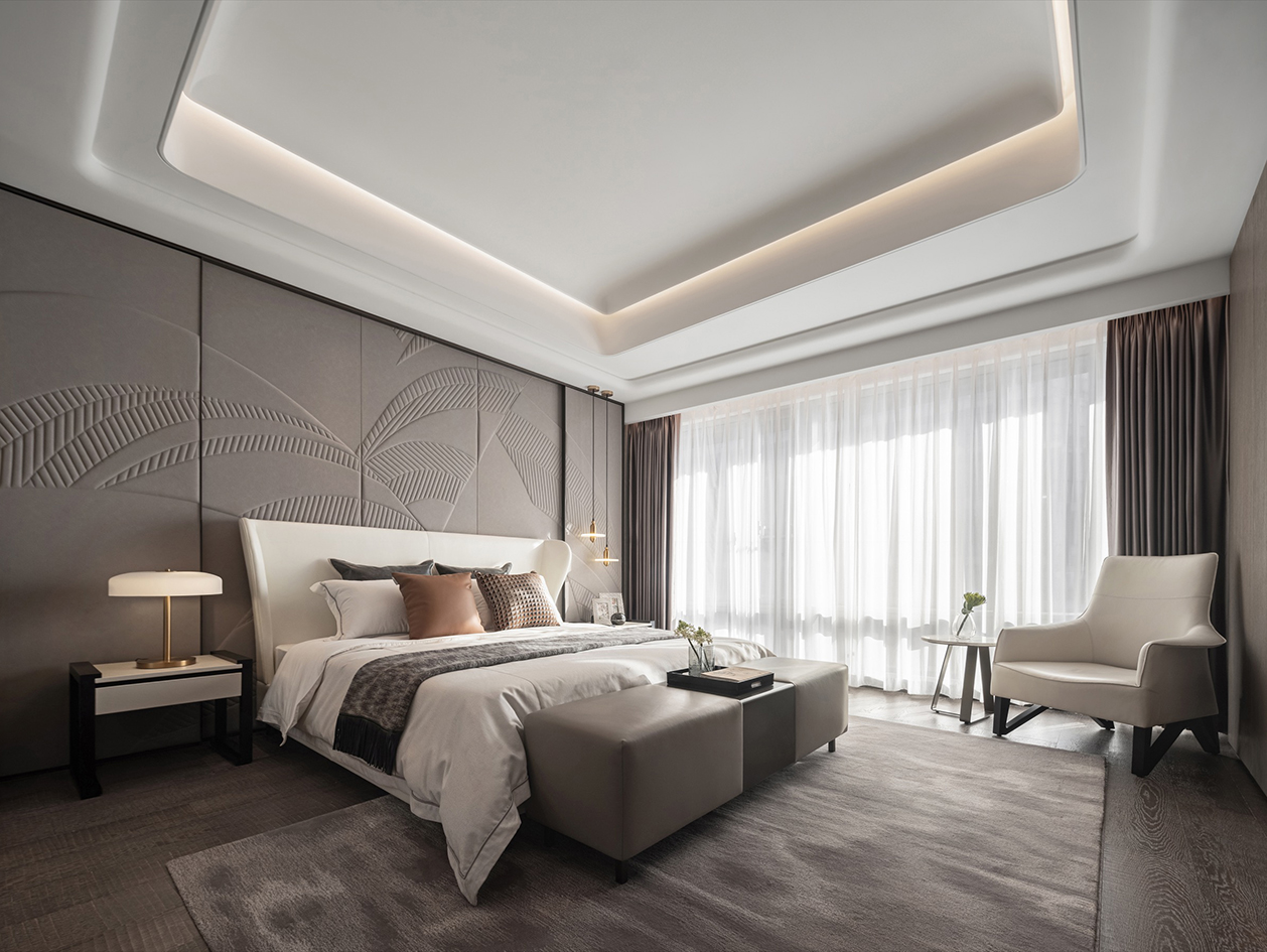Contemporary bedroom with an upholstered feature wall, neutral‑toned bed and bench, soft cove ceiling lighting, bedside lamps, and a lounge chair beside floor‑to‑ceiling sheer curtains