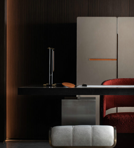 Modern interior workspace with a dark wood wall, minimalist desk, red upholstered chair, two light stools, and a feature stone wall with warm accent lighting.