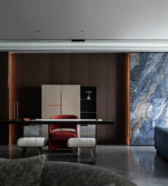 Modern interior workspace with a dark wood wall, minimalist desk, red upholstered chair, two light stools, and a feature stone wall with warm accent lighting.